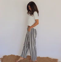 Woman wearing a white top and striped pants standing on a sandy surface with a plain background