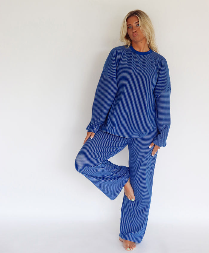 A woman modeling a matching blue striped lounge set consisting of a long-sleeve top and wide-leg pants, standing against a plain white studio background.