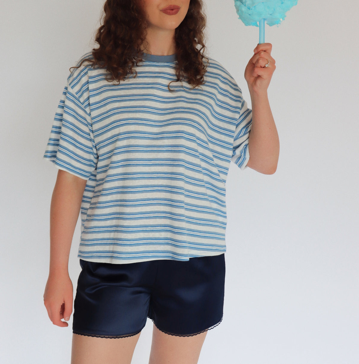 A woman with curly brown hair stands against a plain white background, wearing a loose blue and white striped t-shirt and dark navy shorts, while holding a light blue cotton candy stick.