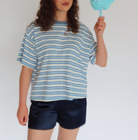A woman with curly brown hair stands against a plain white background, wearing a loose blue and white striped t-shirt and dark navy shorts, while holding a light blue cotton candy stick.
