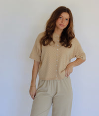 A woman modeling a beige short-sleeve button-front top with a subtle dotted texture, paired with beige and white striped pants, against a plain light background.