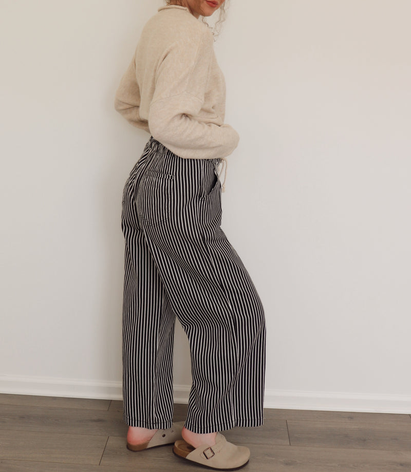 Person wearing a beige sweater and black and white striped pants against a white wall.