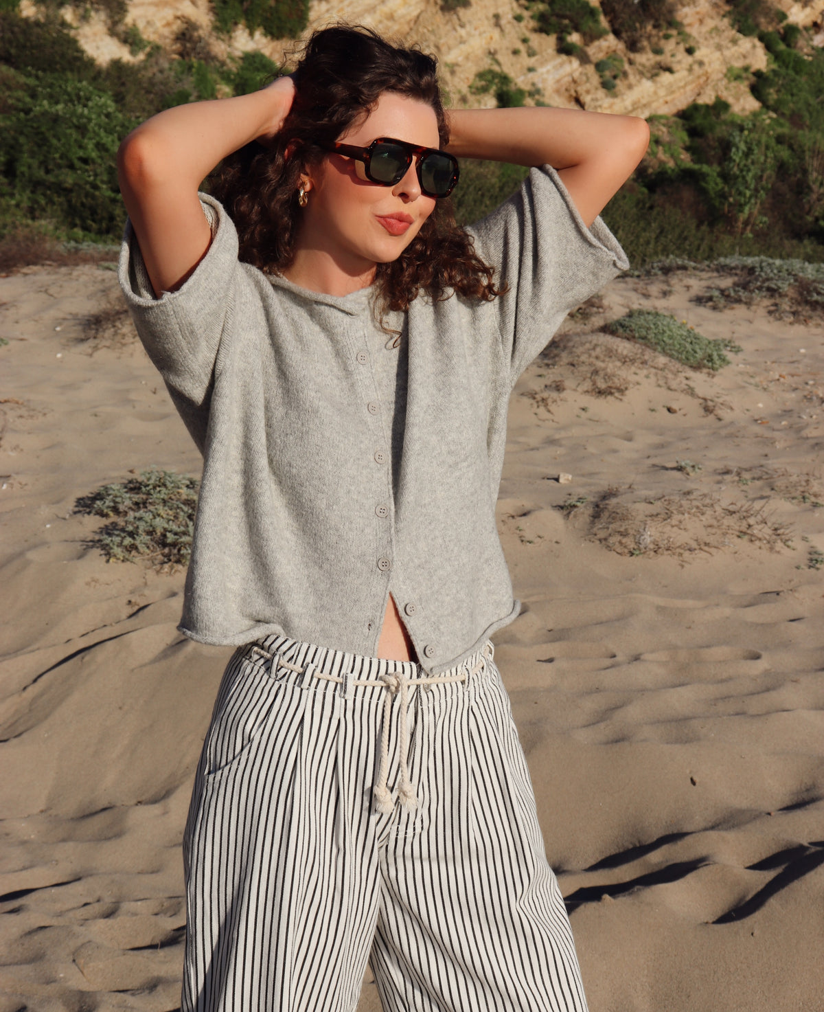 Person wearing a gray top and striped pants standing on a sandy beach.
