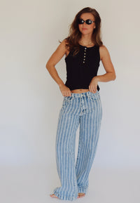A woman modeling a black sleeveless top with star buttons and light blue and white vertically striped wide-leg jeans, standing against a plain white studio background.