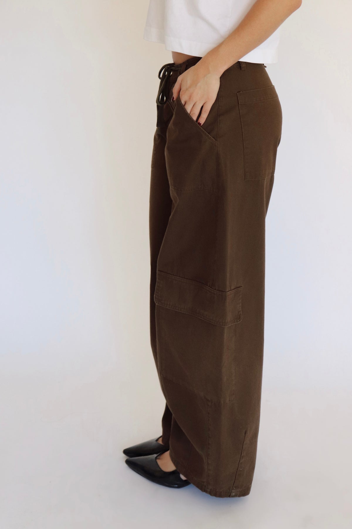 A woman stands against a plain white studio background wearing a white cropped short-sleeve top, loose dark brown wide-leg pants, and black pointed shoes.