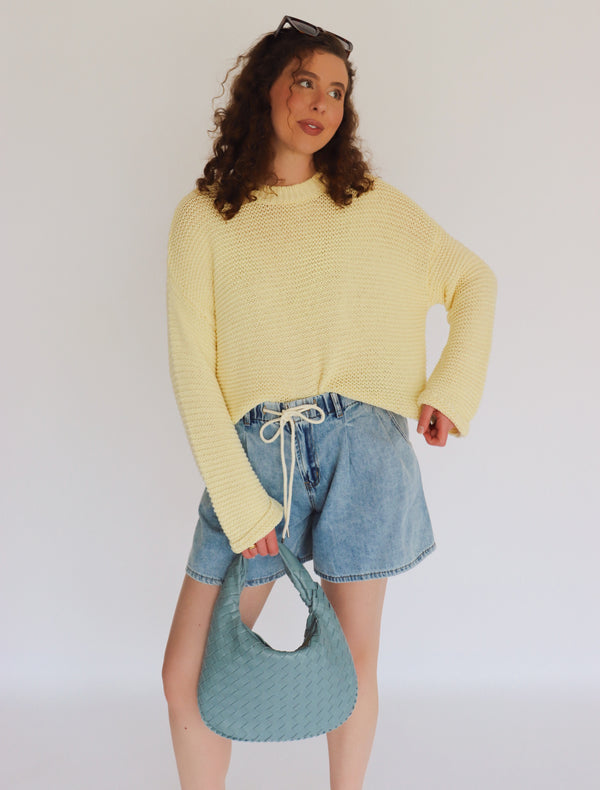 Woman wearing a yellow sweater and denim shorts, holding a blue bag against a white background