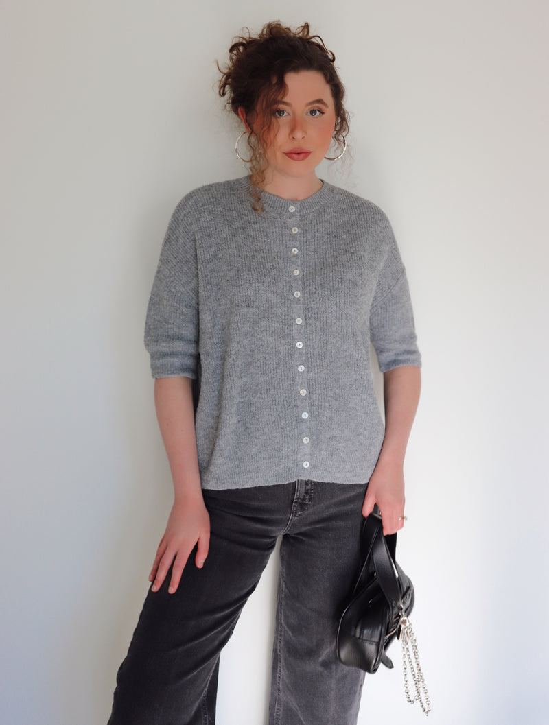 Woman wearing a gray cardigan and black pants against a white wall
