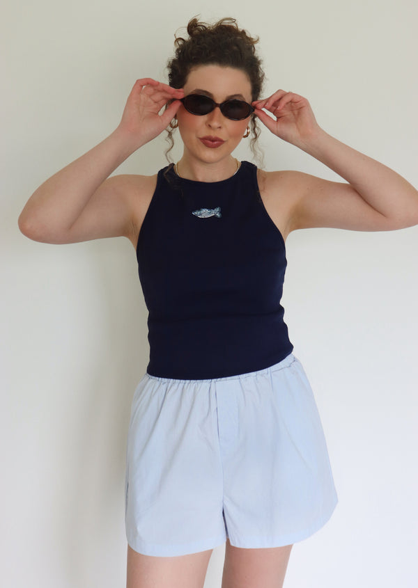 Woman wearing a navy tank top with a fish design and light blue shorts, holding sunglasses.