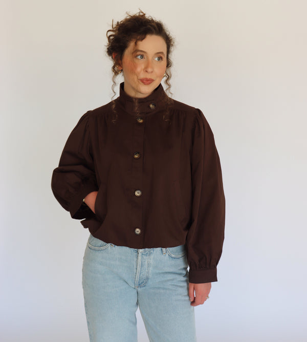 A woman wearing a dark brown long-sleeve button-up blouse jacket with a high mock neck and light blue jeans, standing against a plain white studio background.