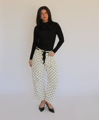 A woman in a black turtleneck and white barrel jeans with black polka dots on them, in front of a white background