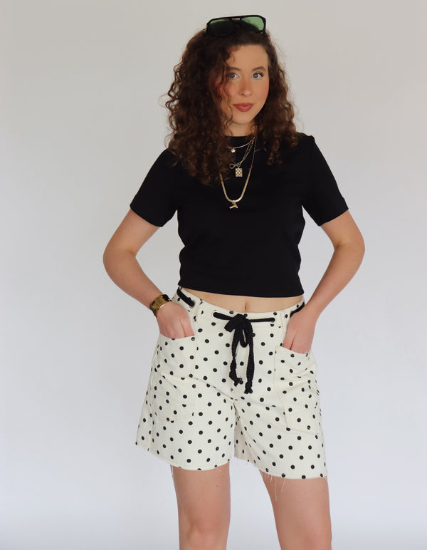 Woman wearing a black crop top and polka dot shorts against a plain background
