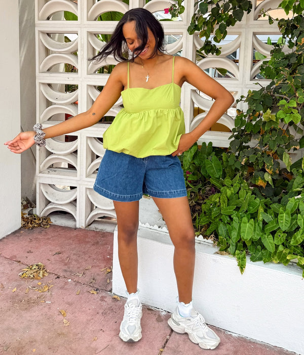 Woman wearing a green top and blue shorts standing outdoors with a decorative white wall and greenery in the background.
