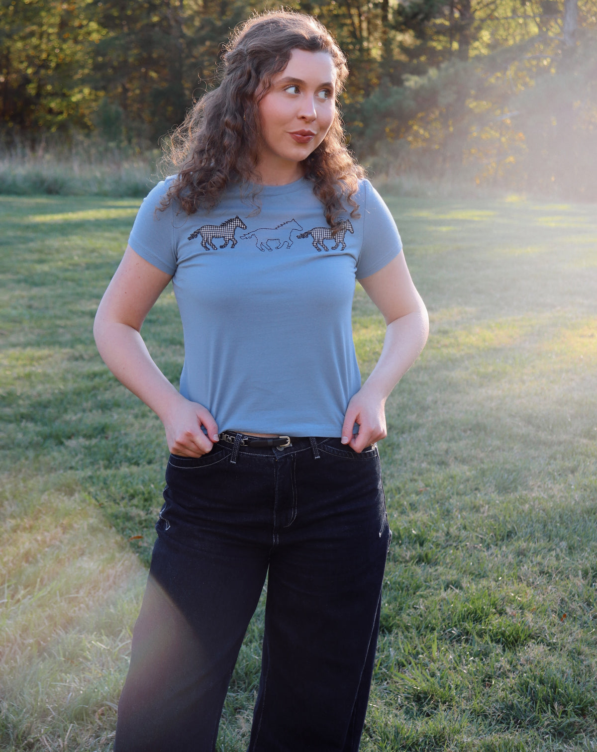 A woman wearing a light blue crewneck t-shirt with a graphic of running horses across the chest, paired with black jeans, standing in a grassy outdoor field during golden hour.