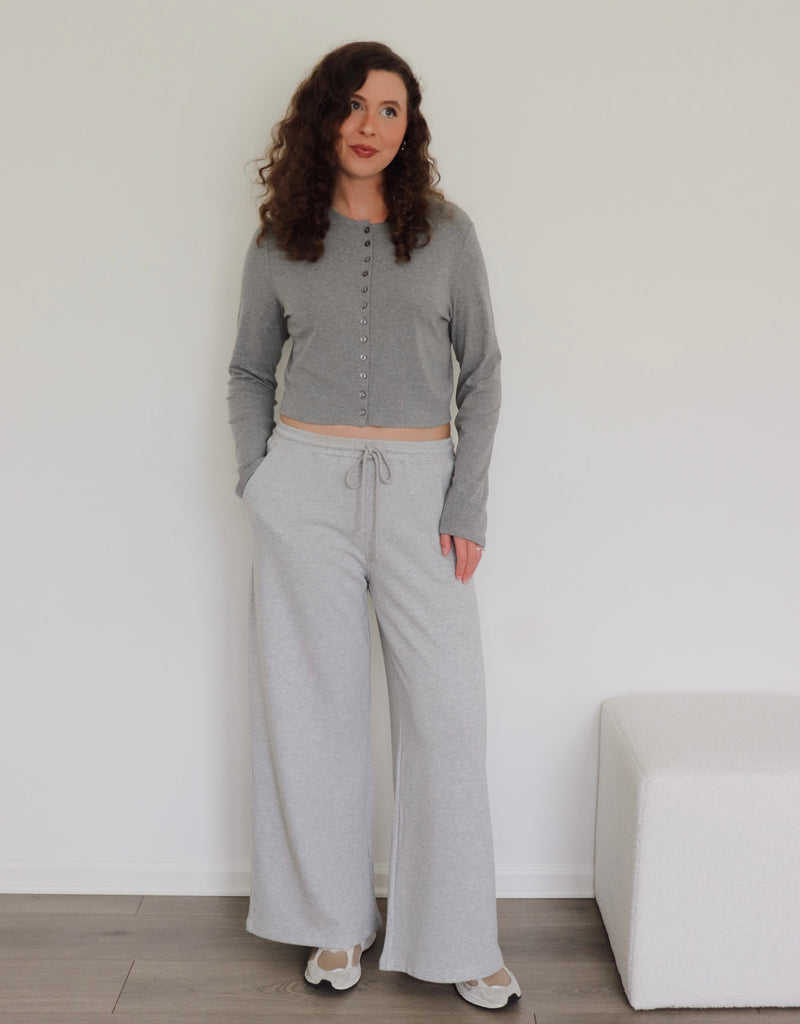 A woman modeling a gray two-piece loungewear outfit consisting of a cropped long-sleeve button-up top and gray wide-leg sweatpants, standing against a white wall.
