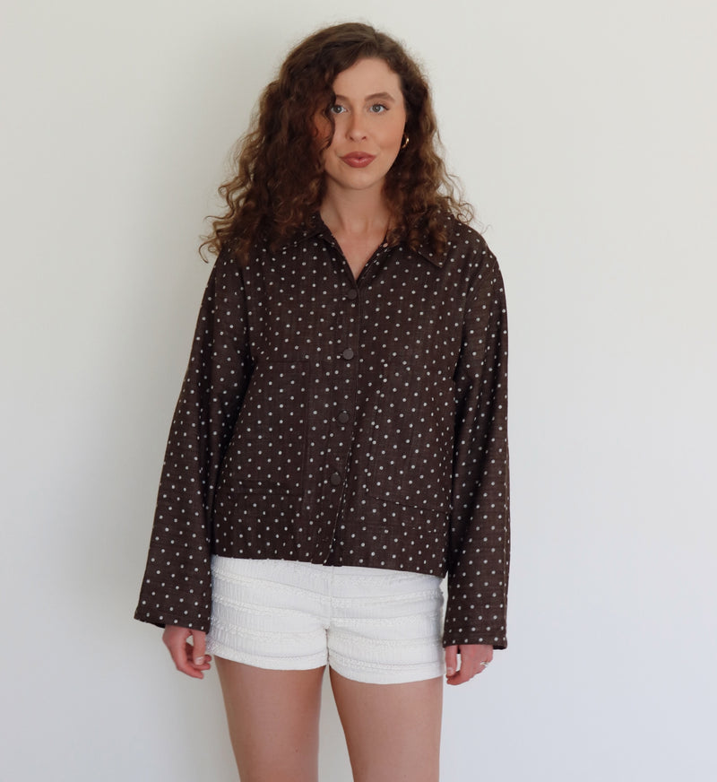 A woman with curly brown hair wearing a dark brown long-sleeve button-up jacker with a white polka-dot pattern and white textured shorts, standing against a plain off-white wall.