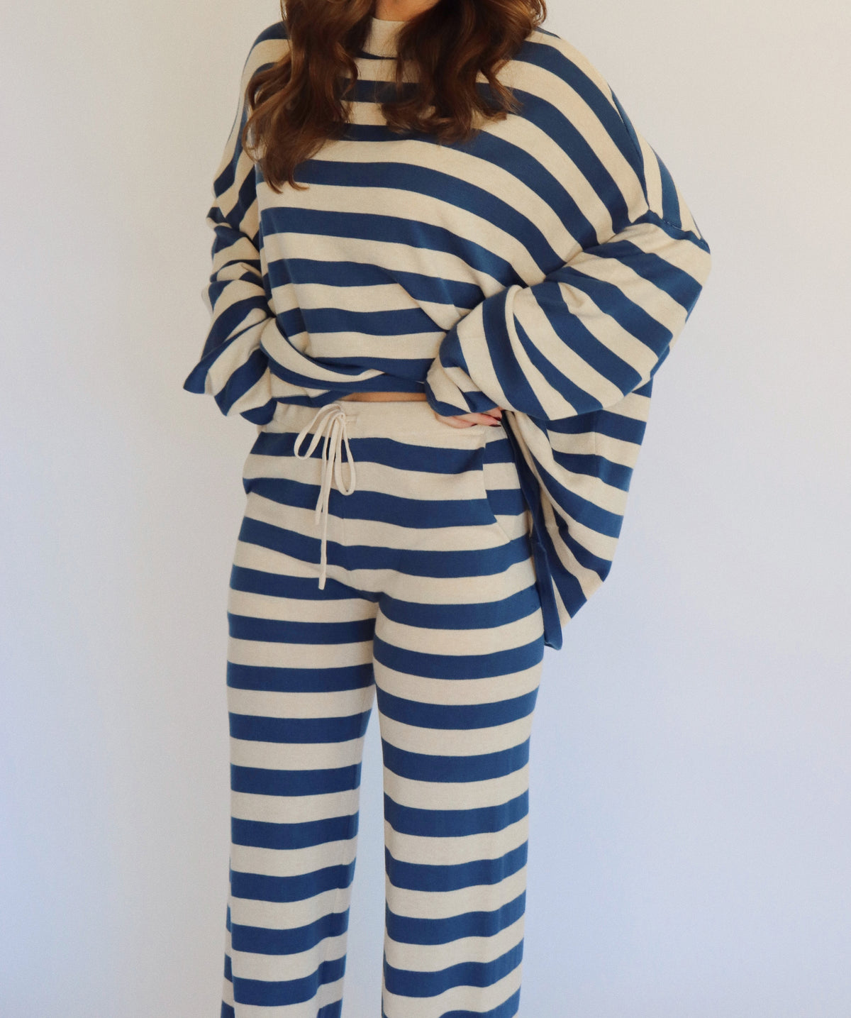 A woman wearing a matching two-piece loungewear set with blue and beige horizontal stripes, featuring a long-sleeve top and wide-leg pants, standing against a plain white background.