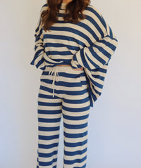 A woman wearing a matching two-piece loungewear set with blue and beige horizontal stripes, featuring a long-sleeve top and wide-leg pants, standing against a plain white background.