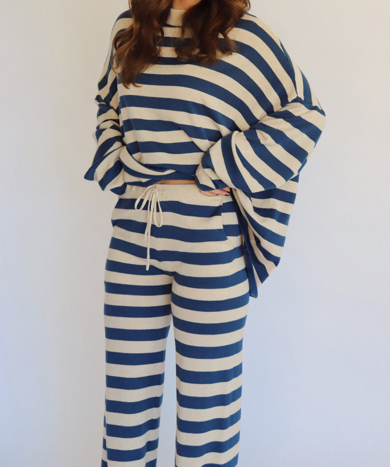 A woman wearing a matching two-piece loungewear set with blue and beige horizontal stripes, featuring a long-sleeve top and wide-leg pants, standing against a plain white background.