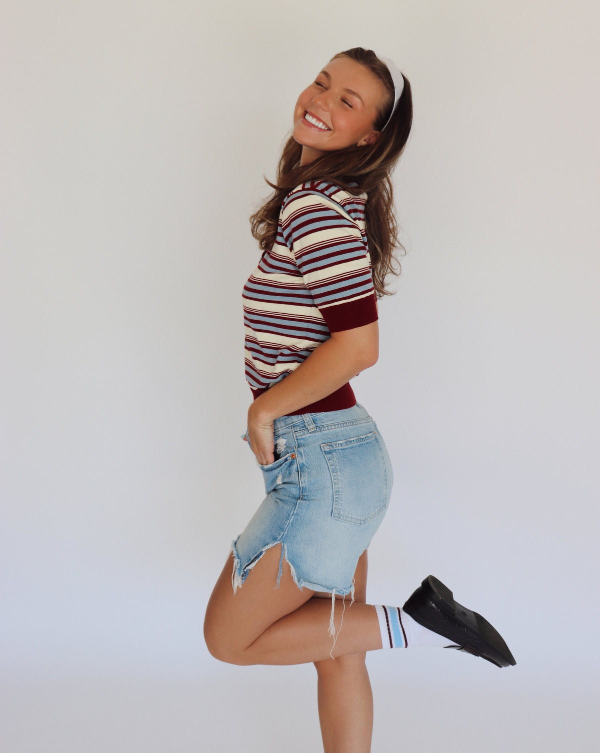 A woman modeling a short-sleeve knit polo shirt with horizontal stripes in cream, burgundy, and blue, paired with light-wash denim shorts.