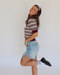 A woman modeling a short-sleeve knit polo shirt with horizontal stripes in cream, burgundy, and blue, paired with light-wash denim shorts.