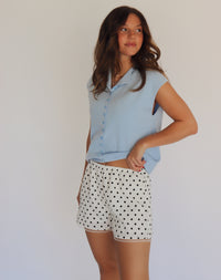 A woman modeling a light blue short-sleeve button-up shirt and white shorts with black polka dots, standing against a plain light background.