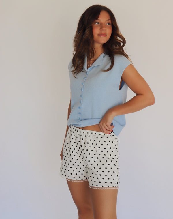 A woman modeling a light blue short-sleeve button-up shirt and white shorts with black polka dots, standing against a plain light background.