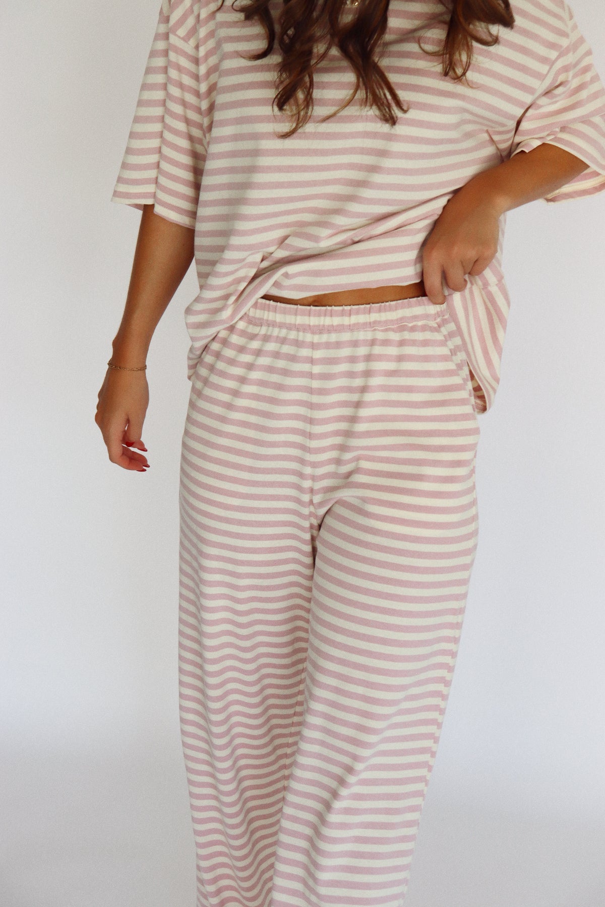 A woman modeling a matching two-piece loungewear set with short-sleeve top and wide-leg pants, featuring horizontal stripes in a light pink and off-white color, set against a plain white studio background.