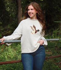 A woman wearing a cream/gray colored knit sweater with a detailed bird motif on the chest, paired with blue jeans, standing outdoors in a wooded setting.