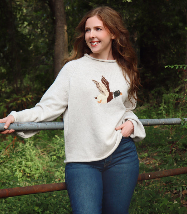 A woman wearing a cream/gray colored knit sweater with a detailed bird motif on the chest, paired with blue jeans, standing outdoors in a wooded setting.