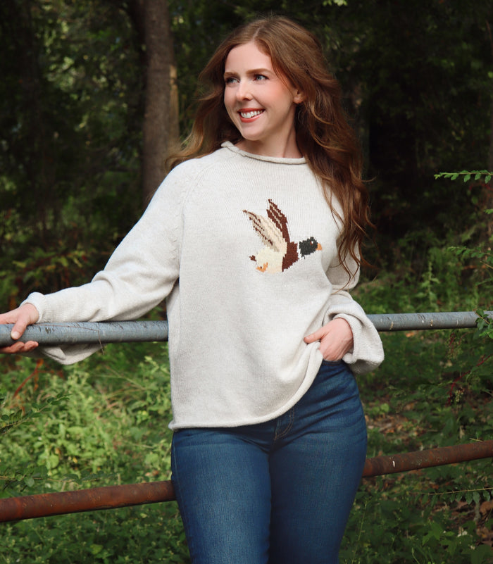 A woman wearing a cream/gray colored knit sweater with a detailed bird motif on the chest, paired with blue jeans, standing outdoors in a wooded setting.