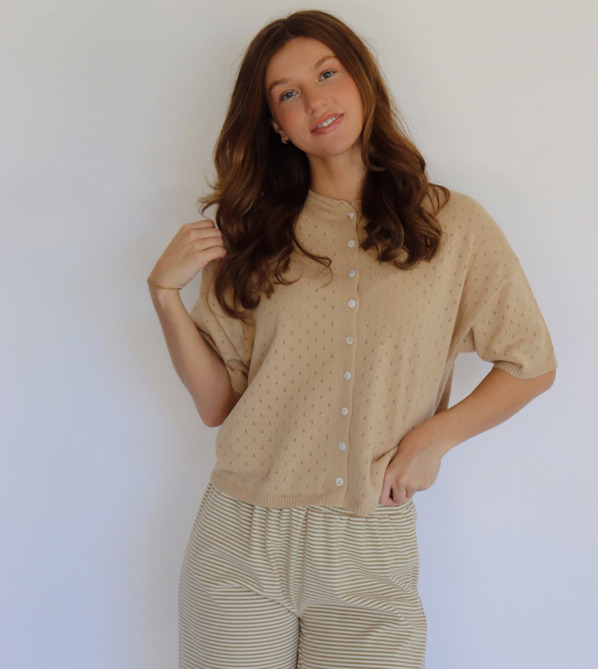 A woman modeling a beige short-sleeve button-front top with a subtle dotted texture, paired with beige and white striped pants, against a plain light background.