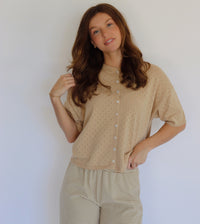 A woman modeling a beige short-sleeve button-front top with a subtle dotted texture, paired with beige and white striped pants, against a plain light background.