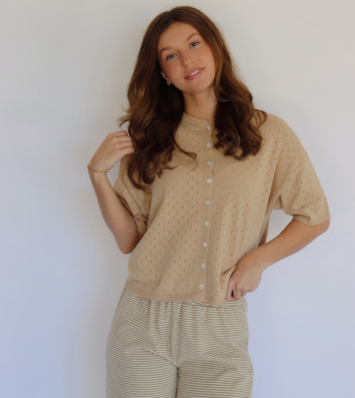A woman modeling a beige short-sleeve button-front top with a subtle dotted texture, paired with beige and white striped pants, against a plain light background.