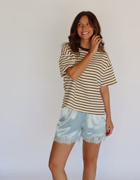 A woman modeling a beige and brown horizontally striped short-sleeve t-shirt with light blue satin shorts featuring a lace hem, standing against a plain white studio background.