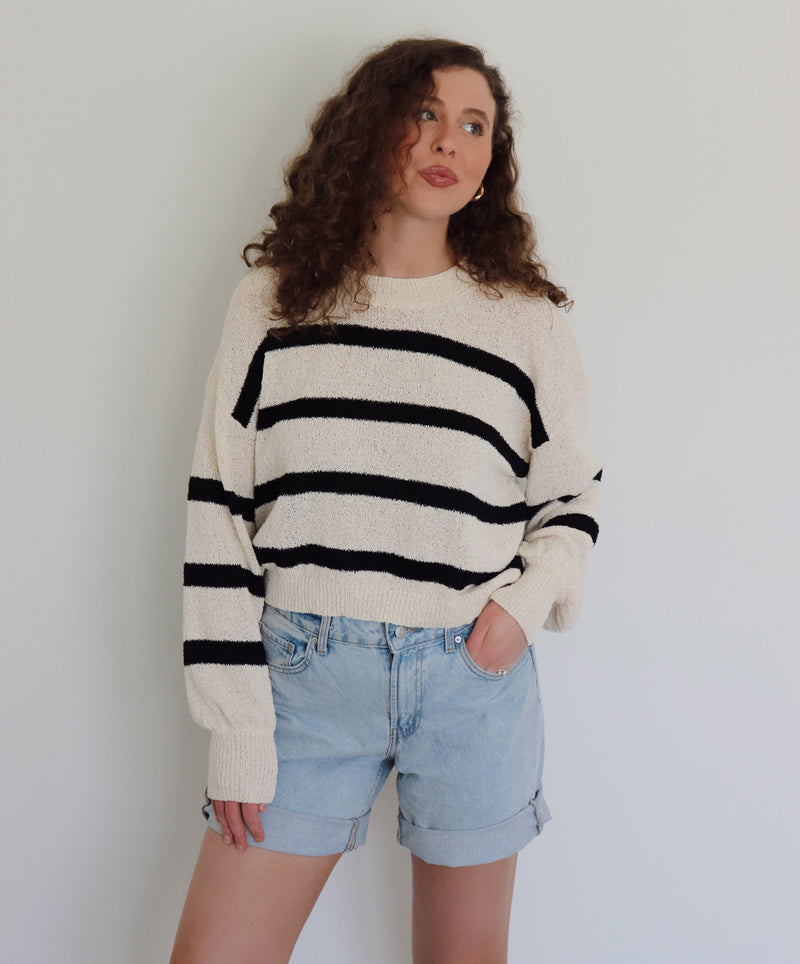 A woman with curly brown hair wearing a cream-colored knit sweater with bold black horizontal stripes and light-wash denim shorts, standing against a plain light gray wall.