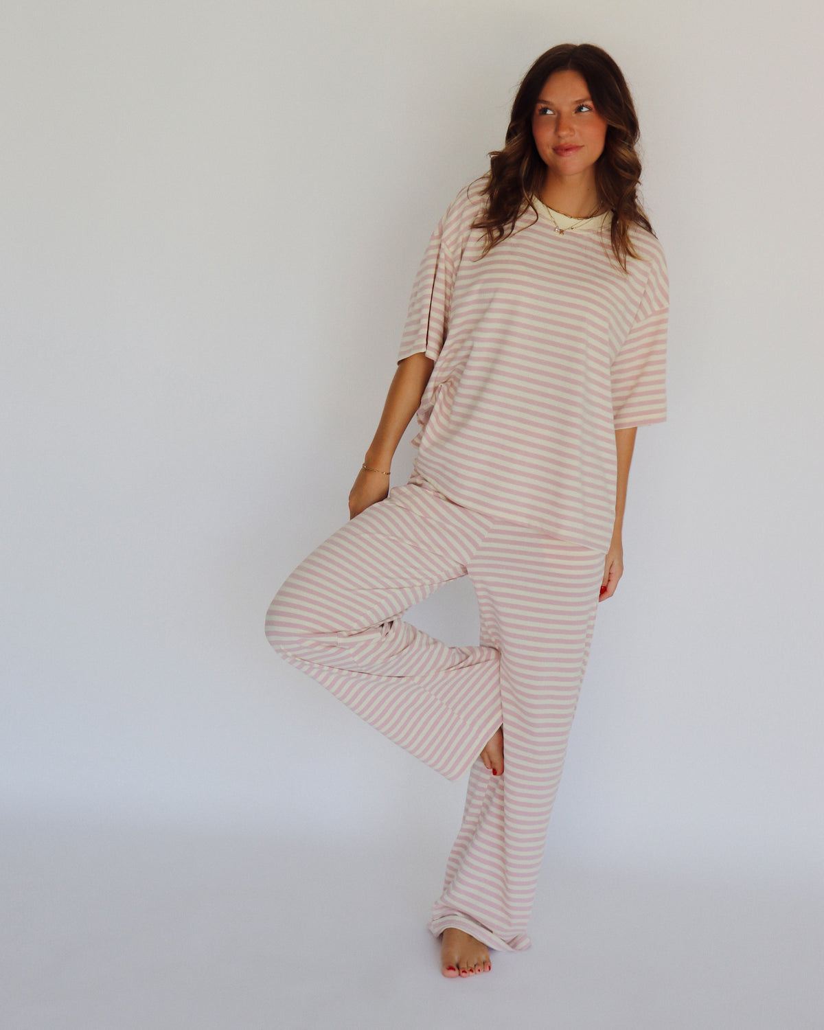 A woman modeling a matching two-piece loungewear set with short-sleeve top and wide-leg pants, featuring horizontal stripes in a light pink and off-white color, set against a plain white studio background.