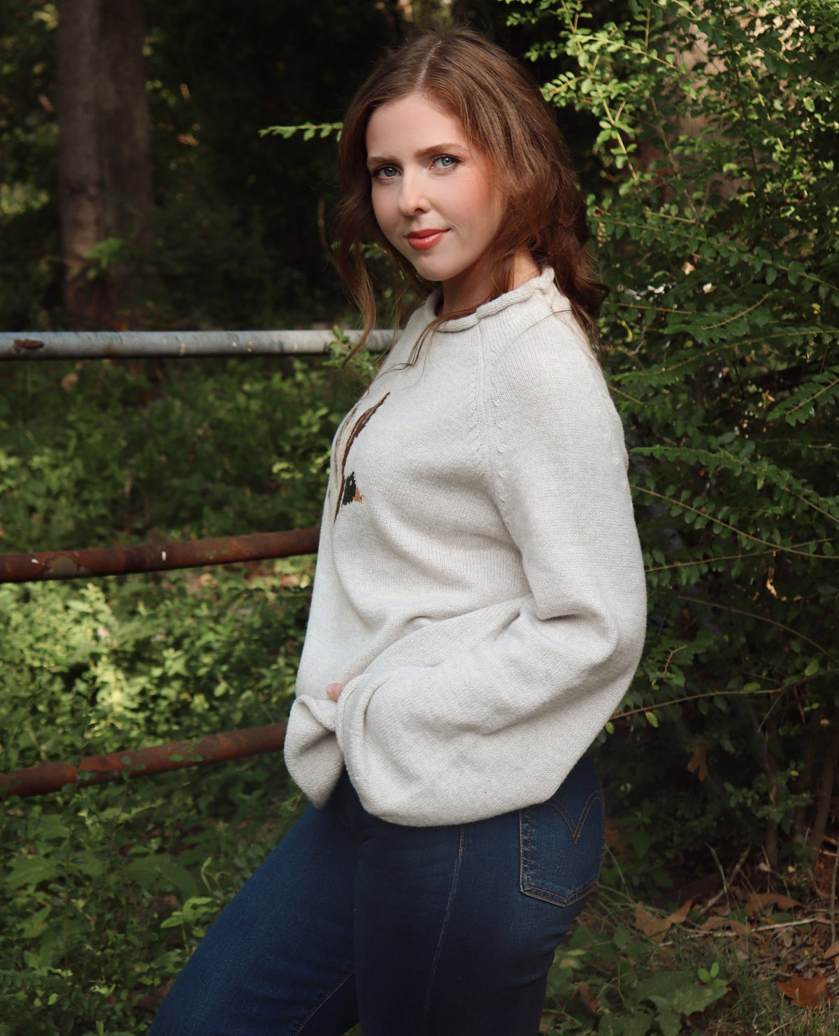 A woman wearing a cream/gray colored knit sweater paired with blue jeans, standing outdoors in a wooded setting.