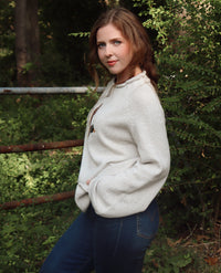 A woman wearing a cream/gray colored knit sweater paired with blue jeans, standing outdoors in a wooded setting.