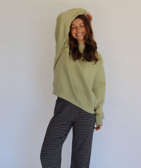 A woman wearing a light sage green oversized mock neck and navy-and-white horizontally striped pants, posing against a plain white wall.