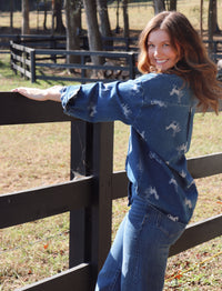 A woman wearing a blue denim shirt with white wild horse prints and matching blue jeans, leaning against a dark wooden fence in an outdoor paddock.