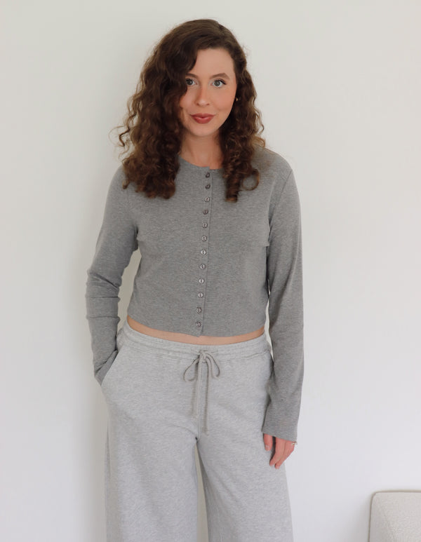 A woman modeling a light gray long-sleeve cropped button-front cardigan with  light gray sweatpants, standing against a plain white wall.