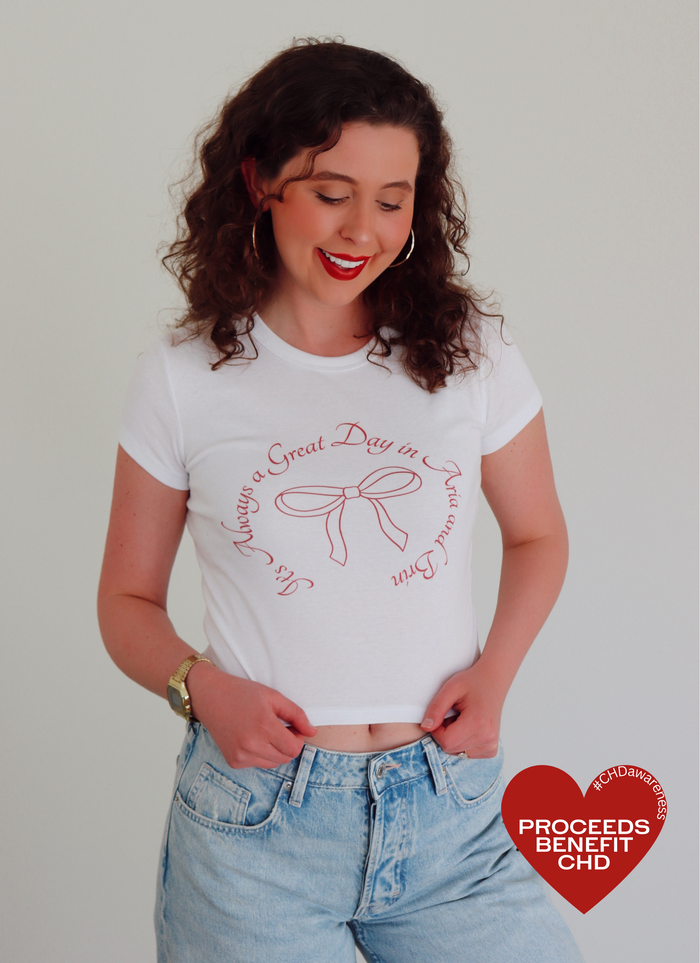 A woman wearing a white crop top with red writing that says "It's Always a Great Day in Aria and Brin" with a bow in the middle 